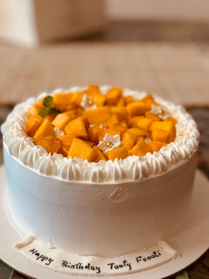 social-feed-img-04 Close-up of a mango-topped cake with white cream frosting and a birthday message.