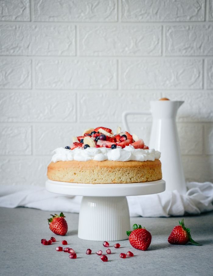 about-img A scrumptious berry-topped cake on a white stand, surrounded by fresh strawberries.